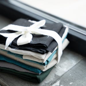 May include: A bundle of folded fabric napkins in shades of black, teal, and green, secured with a white ribbon. The napkins are stacked, indicating a set for table settings or home decoration. The lighting accentuates the textures and colours of the fabric.