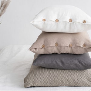 May include: A stack of four linen pillow covers with wooden button closures. The top pillow cover is white, the second is beige, the third is grey, and the bottom is a striped beige and white.