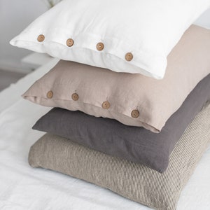 May include: A stack of four linen pillowcases with wooden buttons. The top pillowcase is white, the second is beige, the third is grey, and the bottom is a striped beige and brown.