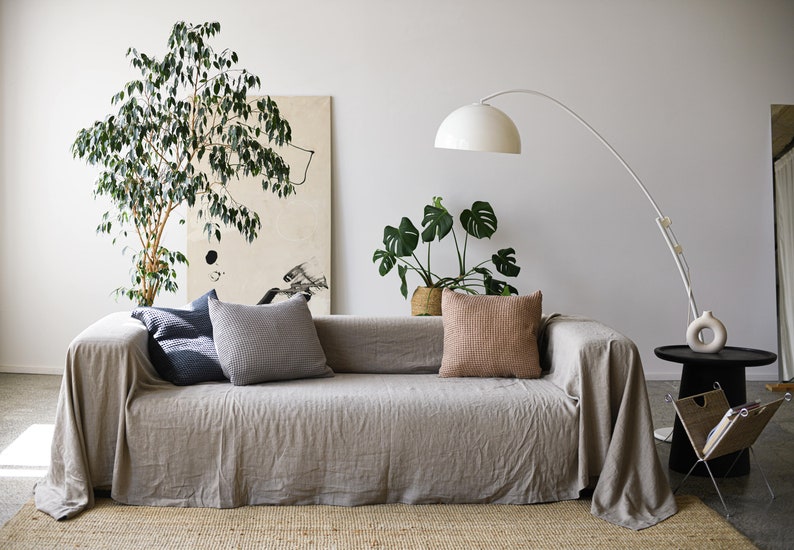 May include: A light gray linen sofa with a throw blanket, three pillows, and two houseplants. The sofa is in a living room with a white wall and a floor lamp.