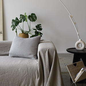 May include: A light gray linen throw blanket draped over a couch with a white and gray patterned pillow. A white arc lamp with a white shade is in the background.