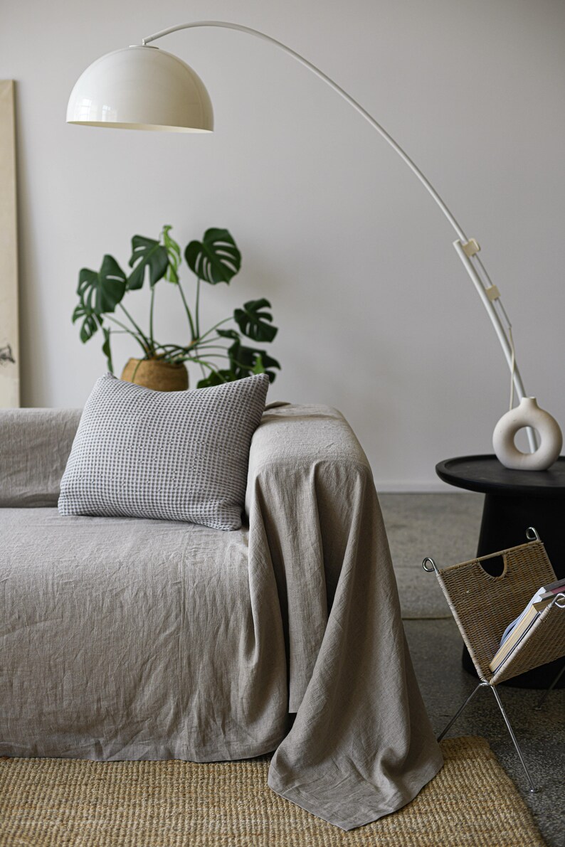 May include: A light gray linen throw blanket draped over a couch with a white and gray patterned pillow. A white arc lamp with a white shade is in the background.