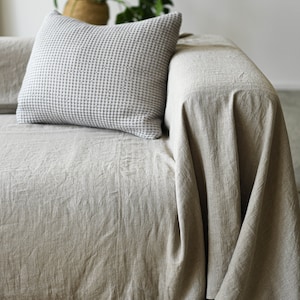 May include: A light beige linen throw draped over a couch with a white and gray checkered pillow. The couch is sitting on a woven rug.