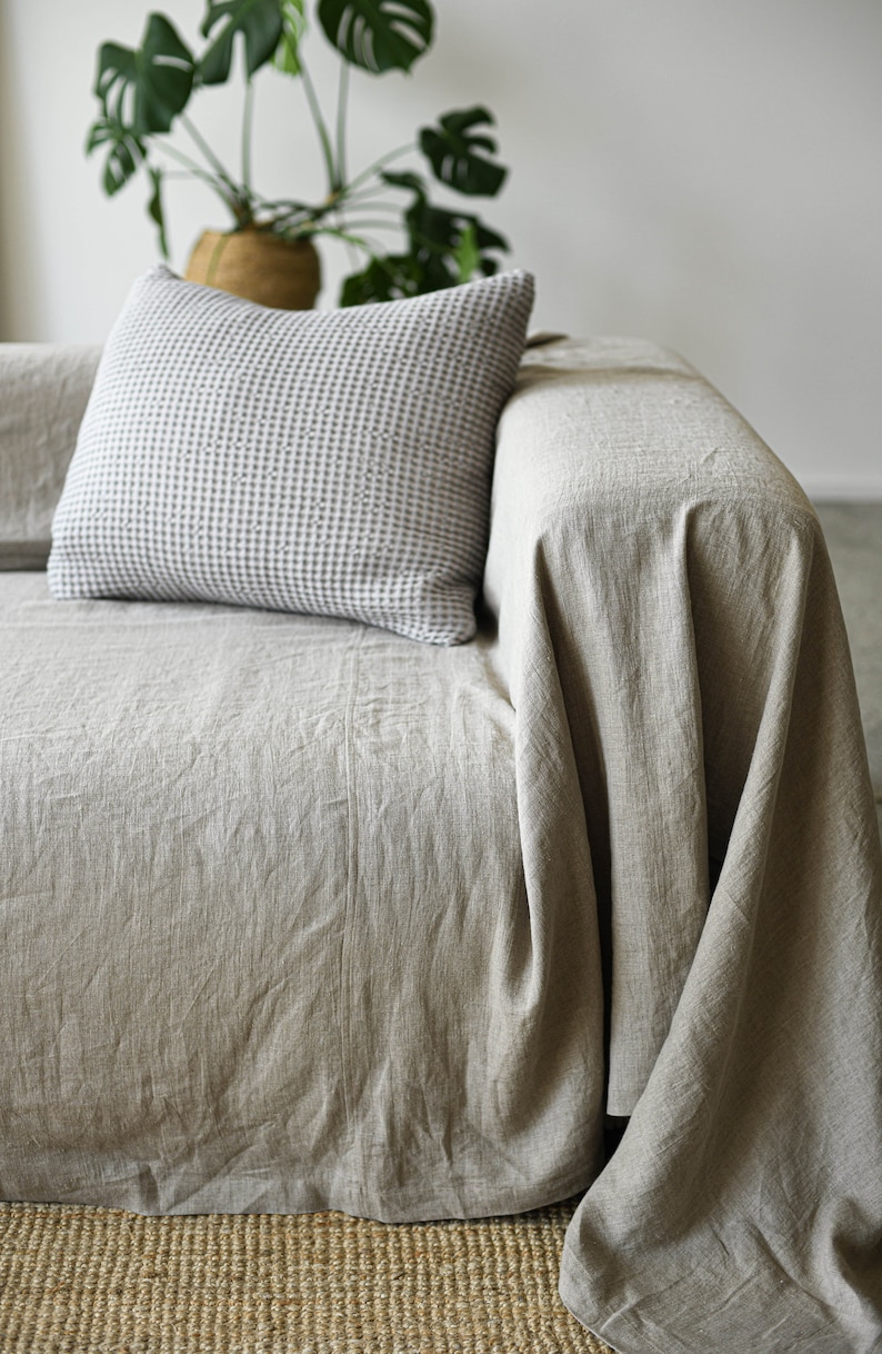 May include: A light beige linen throw draped over a couch with a white and gray checkered pillow. The couch is sitting on a woven rug.