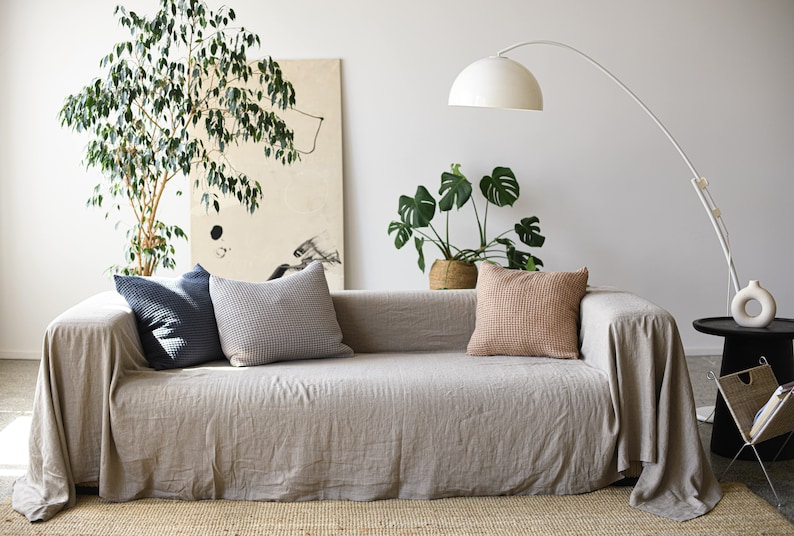 May include: A light gray linen sofa with a throw blanket draped over it. The sofa has three pillows, one dark blue, one gray and white checkered, and one light brown. There are two plants in the background, one with large green leaves and one with small green leaves. A white floor lamp with a curved arm is in the background.