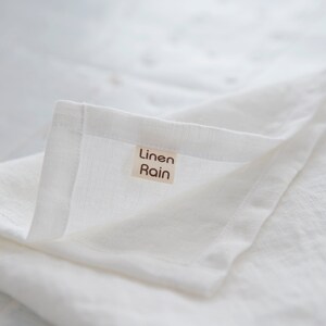 May include: Close-up of a folded white linen cloth. The fabric has a slightly textured appearance. A small rectangular tag with the words "Linen Rain" is sewn onto the corner of the cloth. The overall aesthetic is clean and minimalist.