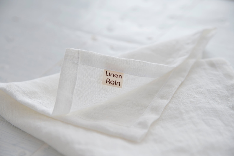 May include: Close-up of a folded white linen cloth. The fabric has a slightly textured appearance. A small rectangular tag with the words "Linen Rain" is sewn onto the corner of the cloth. The overall aesthetic is clean and minimalist.
