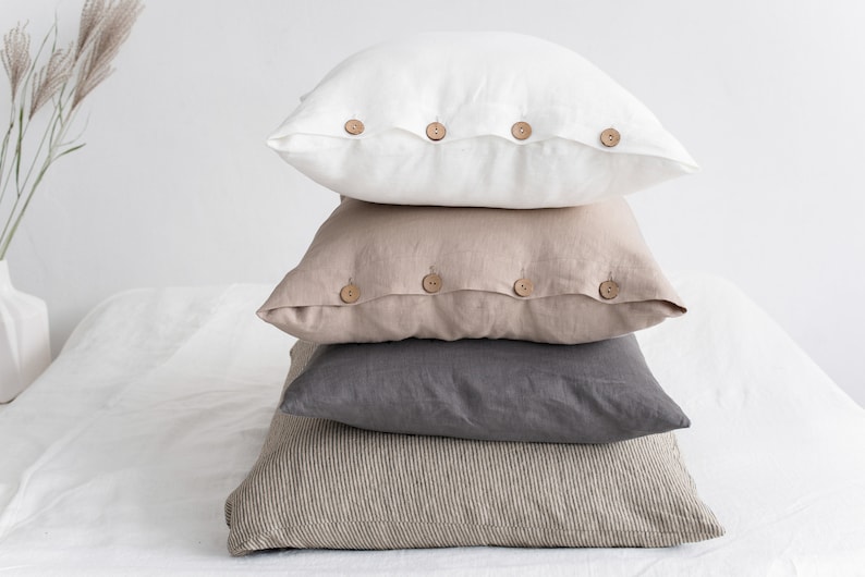 May include: A stack of four linen pillow covers with wooden button closures. The top pillow cover is white, the second is beige, the third is grey, and the bottom is a striped beige and white.
