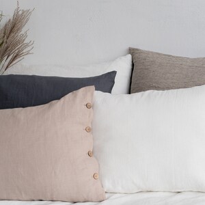 May include: Four decorative throw pillows with button accents. The pillows are arranged in a stack with a beige pillow on the bottom, a white pillow in the middle, a dark grey pillow on top of the white pillow, and a striped brown pillow on top of the grey pillow.