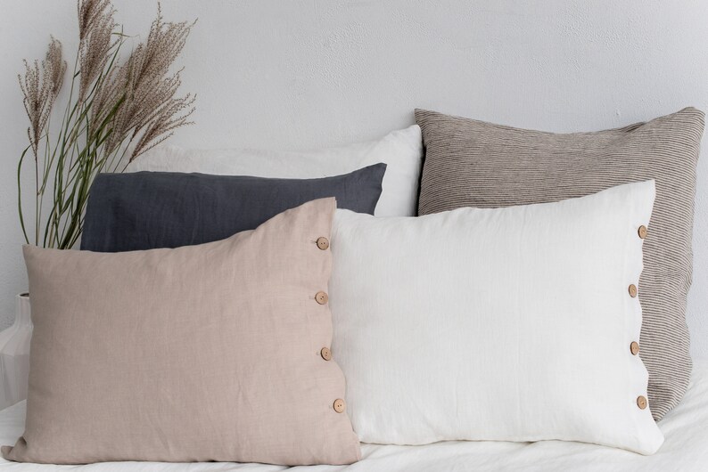 May include: Four decorative throw pillows with button accents. The pillows are arranged in a stack with a beige pillow on the bottom, a white pillow in the middle, a dark grey pillow on top of the white pillow, and a striped brown pillow on top of the grey pillow.