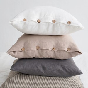 May include: A stack of four linen pillow covers with wooden button closures. The top pillow cover is white, the second is beige, the third is grey, and the bottom is a striped beige and brown.