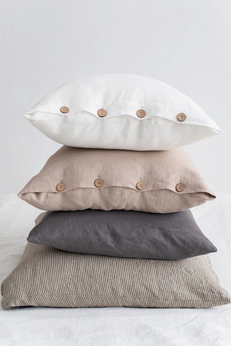 May include: A stack of four linen pillow covers with wooden button closures. The top pillow cover is white, the second is beige, the third is grey, and the bottom is a striped beige and brown.