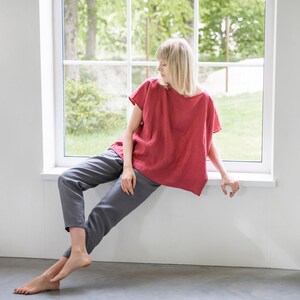 May include: A woman sits by a window wearing a red linen top and gray linen pants.