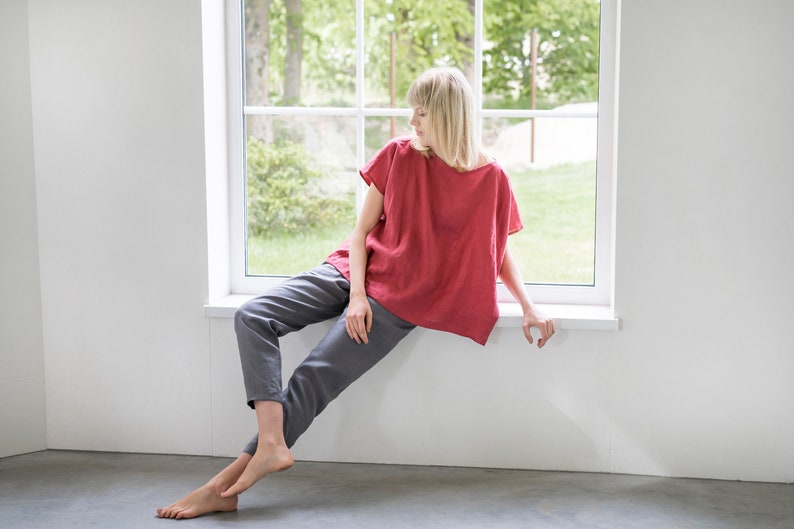 May include: A woman sits by a window wearing a red linen top and gray linen pants.