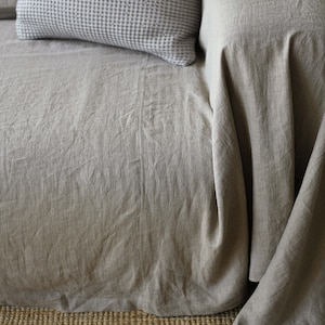 May include: A close-up of a light brown linen sofa with a gray and white patterned pillow. The sofa is sitting on a woven rug.