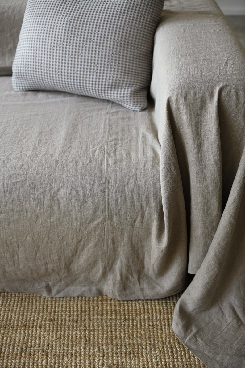 May include: A close-up of a light brown linen sofa with a gray and white patterned pillow. The sofa is sitting on a woven rug.