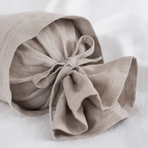 May include: A beige linen bolster pillow with a tied bow at the end. The pillow is made of natural linen fabric and has a simple, elegant design.