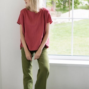 May include: A woman wearing a loose-fitting, coral-colored linen top with short sleeves and olive green linen pants. She is standing near a window with a view of green trees and grass.