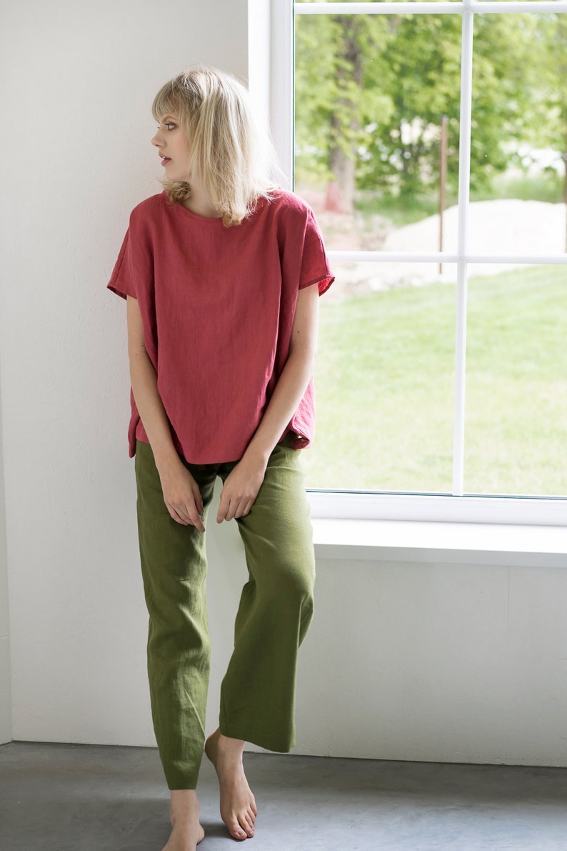 May include: A woman wearing a loose-fitting, coral-colored linen top with short sleeves and olive green linen pants. She is standing near a window with a view of green trees and grass.