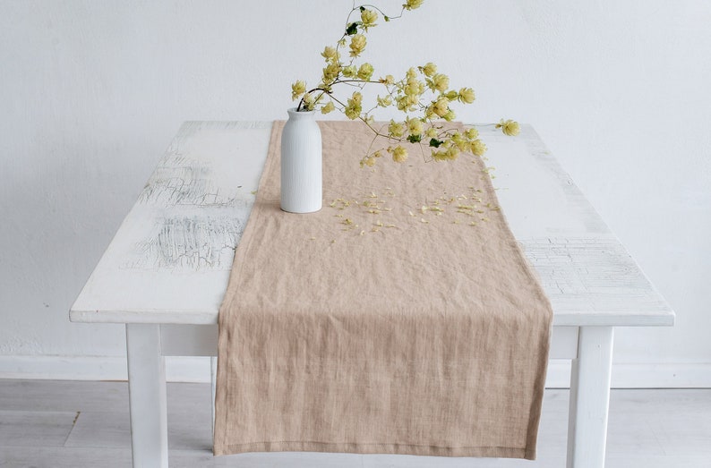 May include: A beige linen table runner draped over a white distressed wooden table. A white ceramic vase holds a sprig of yellow-green flowers. The scene is set against a white wall.