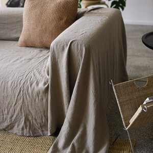 May include: A light brown linen throw blanket draped over a beige sofa with a brown waffle-weave throw pillow. The sofa is in front of a woven basket and a potted plant.
