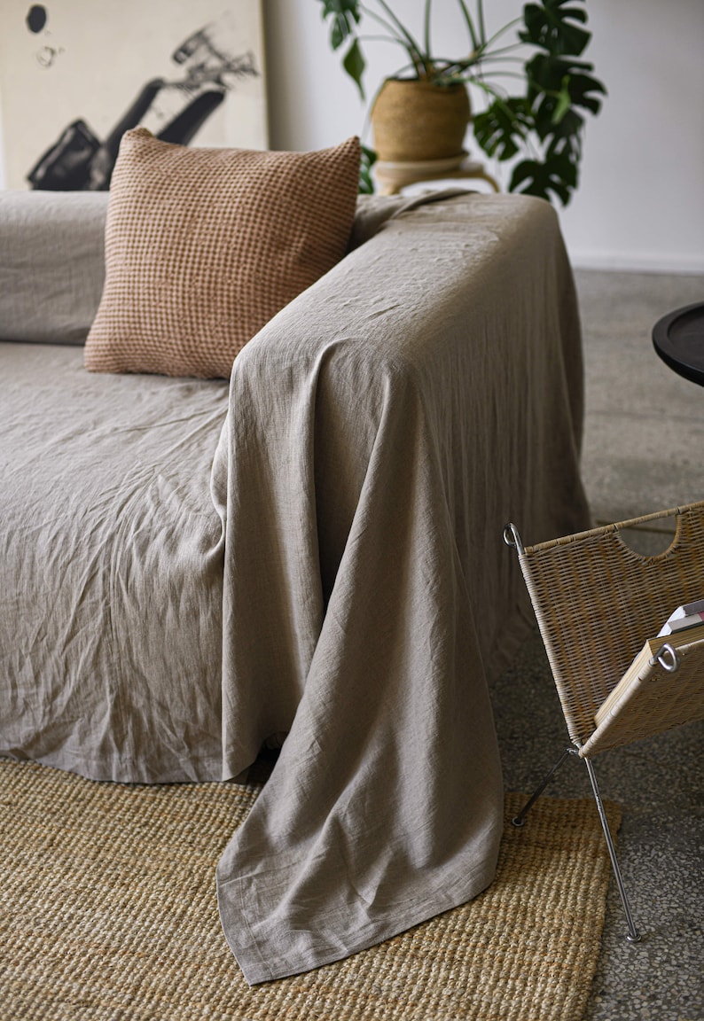 May include: A light brown linen throw blanket draped over a beige sofa with a brown waffle-weave throw pillow. The sofa is in front of a woven basket and a potted plant.