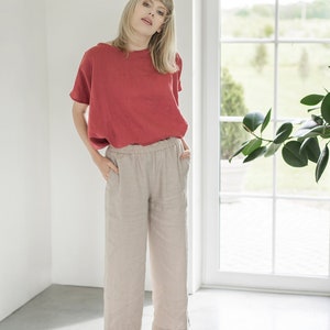 May include: A woman wearing a red linen top and beige linen pants with an elastic waistband. She is standing in a room with a window behind her.