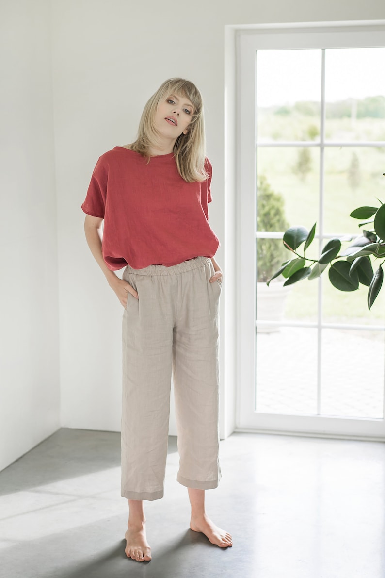 May include: A woman wearing a red linen top and beige linen pants with an elastic waistband. She is standing in a room with a window behind her.