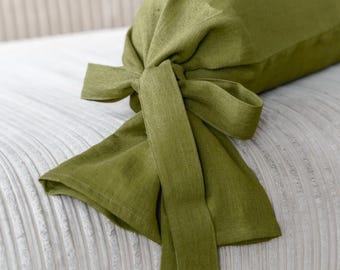 May include: A green bolster pillow with a tied bow detail. The pillow is made of a textured fabric and rests on a cream-colored, ribbed surface. The olive green color adds a touch of nature to the decor.