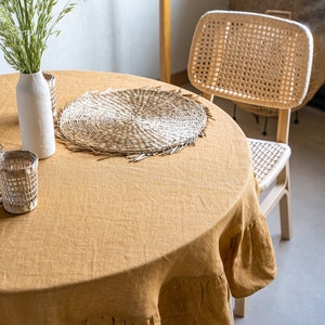 Pu&ograve; includere: Un tovaglia rotonda di lino marrone con un bordo arricciato, un sottopiatto intrecciato e un vaso di vetro trasparente con fiori secchi su un tavolo.