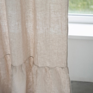 Puede incluir: Primer plano de una cortina de lino beige con un dobladillo con volantes. La cortina est&aacute; colgada delante de una ventana.