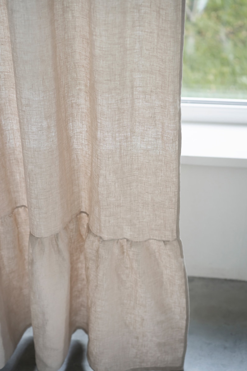 Puede incluir: Primer plano de una cortina de lino beige con un dobladillo con volantes. La cortina est&aacute; colgada delante de una ventana.
