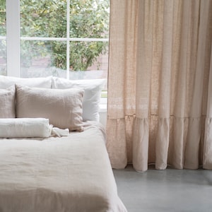 Puede incluir: Una cama con una falda de cama blanca y un edred&oacute;n y almohadas de lino marr&oacute;n claro. La cama est&aacute; junto a una ventana con una cortina de lino marr&oacute;n claro.