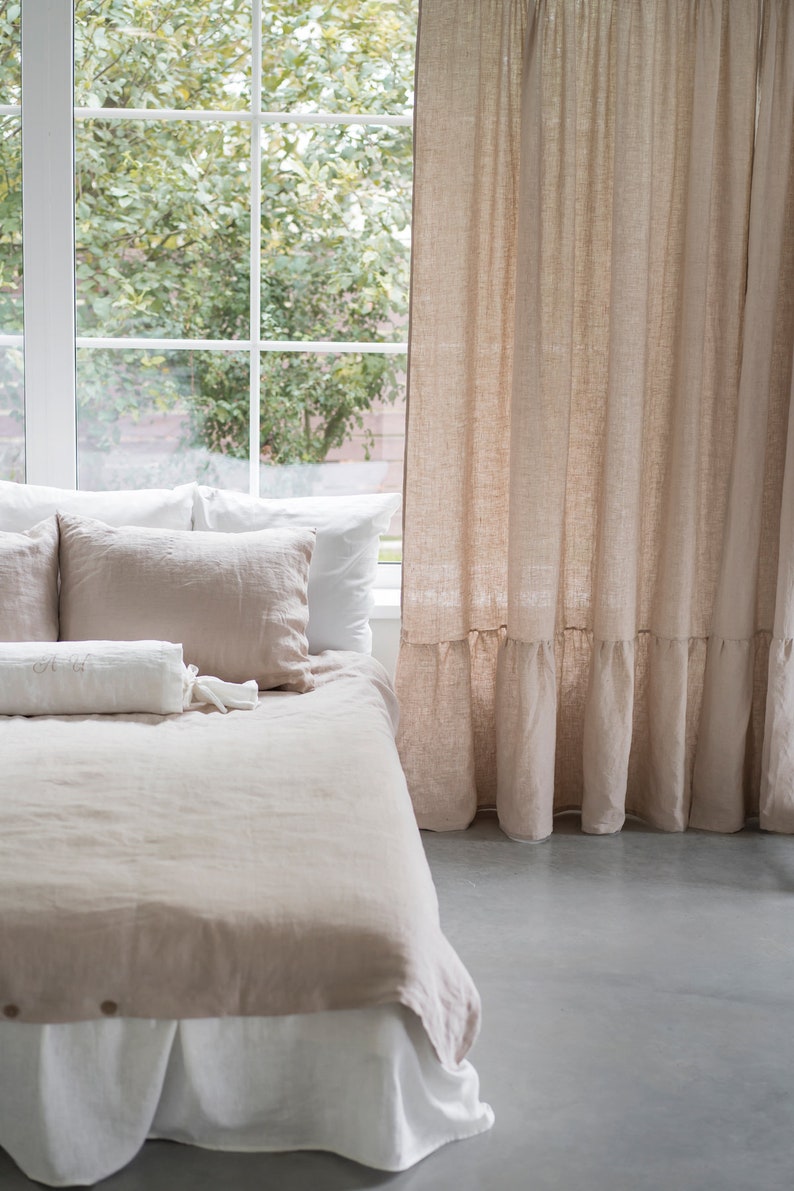 Puede incluir: Una cama con una falda de cama blanca y un edred&oacute;n y almohadas de lino marr&oacute;n claro. La cama est&aacute; junto a una ventana con una cortina de lino marr&oacute;n claro.