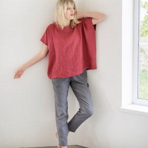 May include: A woman wearing a loose-fitting, red linen top with short sleeves and gray linen pants. She is standing in a corner of a room with a white wall and a window.