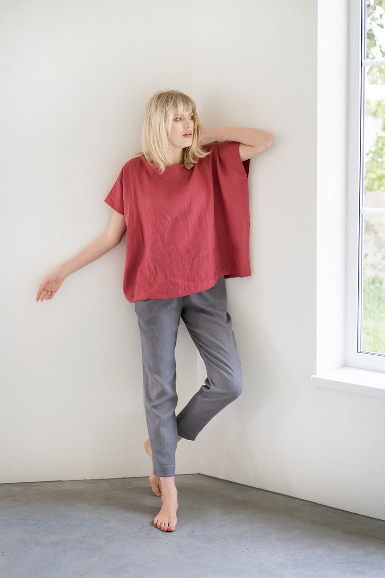 May include: A woman wearing a loose-fitting, red linen top with short sleeves and gray linen pants. She is standing in a corner of a room with a white wall and a window.