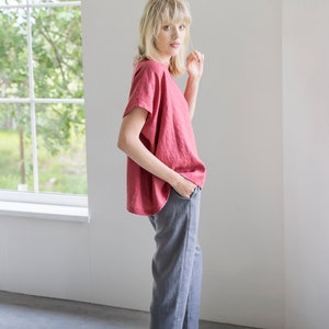 May include: A woman wearing a loose-fitting, coral-colored linen top with short sleeves and gray linen pants. She is standing in a room with a window in the background.