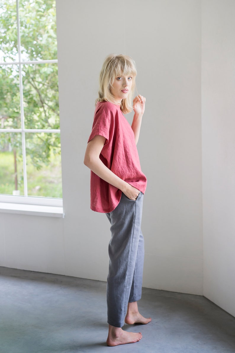 May include: A woman wearing a loose-fitting, coral-colored linen top with short sleeves and gray linen pants. She is standing in a room with a window in the background.