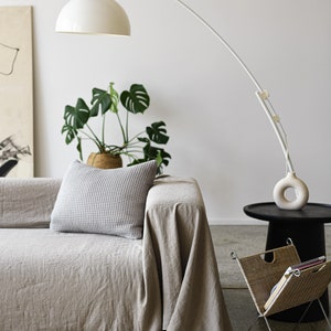 May include: A light gray linen sofa with a white throw blanket and a patterned pillow. A white arched floor lamp with a white shade is in the background. A potted plant is on the floor next to the sofa. A small black side table with a wicker magazine rack is in the foreground.