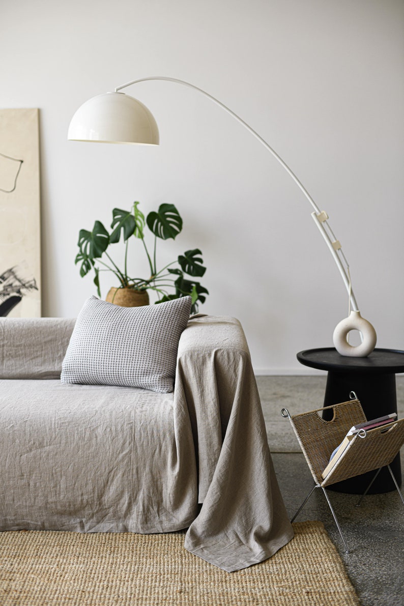May include: A light gray linen sofa with a white throw blanket and a patterned pillow. A white arched floor lamp with a white shade is in the background. A potted plant is on the floor next to the sofa. A small black side table with a wicker magazine rack is in the foreground.
