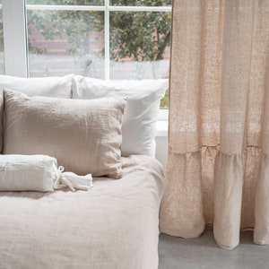 Puede incluir: Una cama con un juego de ropa de cama de lino blanco y beige. La cama tiene una almohada de apoyo blanca con las iniciales "A & H" bordadas. La cama est&aacute; junto a una ventana con una cortina de lino beige con un dobladillo con volantes.