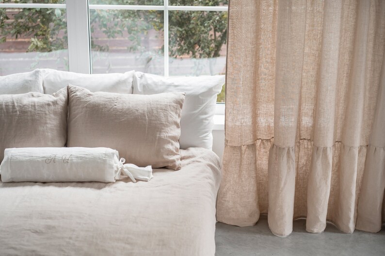 Puede incluir: Una cama con un juego de ropa de cama de lino blanco y beige. La cama tiene una almohada de apoyo blanca con las iniciales "A & H" bordadas. La cama est&aacute; junto a una ventana con una cortina de lino beige con un dobladillo con volantes.