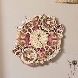 Puede incluir: Un reloj de madera con un diseño complejo de engranajes en tonos rosa y beige. La esfera del reloj muestra números romanos y signos del zodiaco. El reloj está montado en una pared clara.