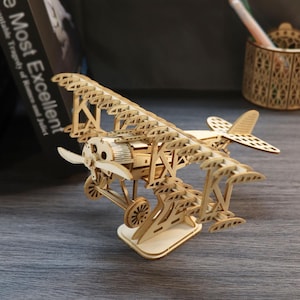 May include: A light-colored wooden model biplane featuring a detailed propeller, two sets of stacked wings, and a landing gear assembly. The structure is composed of laser-cut wooden pieces with visible joints and supports, mounted on a base.