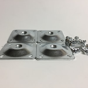 May include: Four silver square furniture leg mounting plates with a central threaded insert and screw holes. A pile of silver screws is next to the plates. Hardware for furniture assembly.