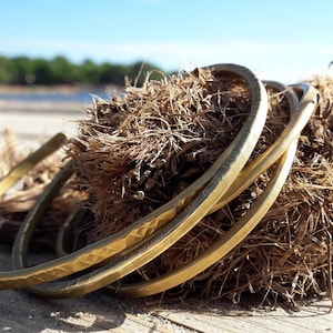 May include: Three gold-toned metal bracelets stacked on top of each other. The bracelets are lying on a brown, fibrous surface.