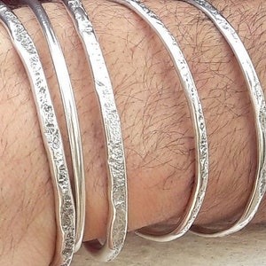 Puede incluir: Cinco pulseras de plata apiladas en la muñeca de una persona. Las pulseras son todas lisas y tienen una textura martillada.