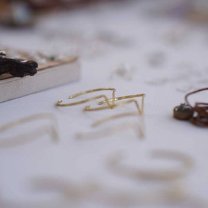 Puede incluir: Un conjunto de tres pulseras de metal dorado con un diseño geométrico. Las pulseras están dispuestas sobre una superficie blanca.