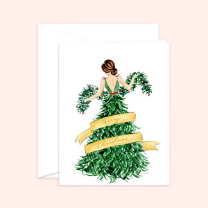 Op de afbeelding: Een aquarel-illustratie van een vrouw die een kerstboomjurk draagt. Ze houdt een krans in elke hand. De jurk heeft een gouden lint eromheen met de woorden "Merry Christmas" erop.