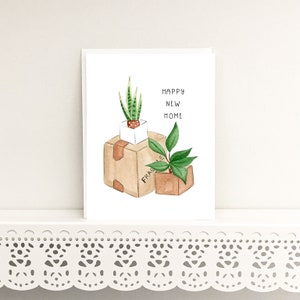Peut inclure: Une carte blanche avec une illustration aquarelle de deux plantes en pot dans des bo&icirc;tes en carton. Le texte "Happy New Home" est &eacute;crit en lettres noires.